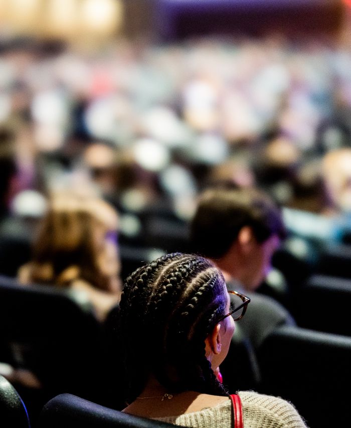 A diverse audience attentively listens to a speaker in a large auditorium setting.